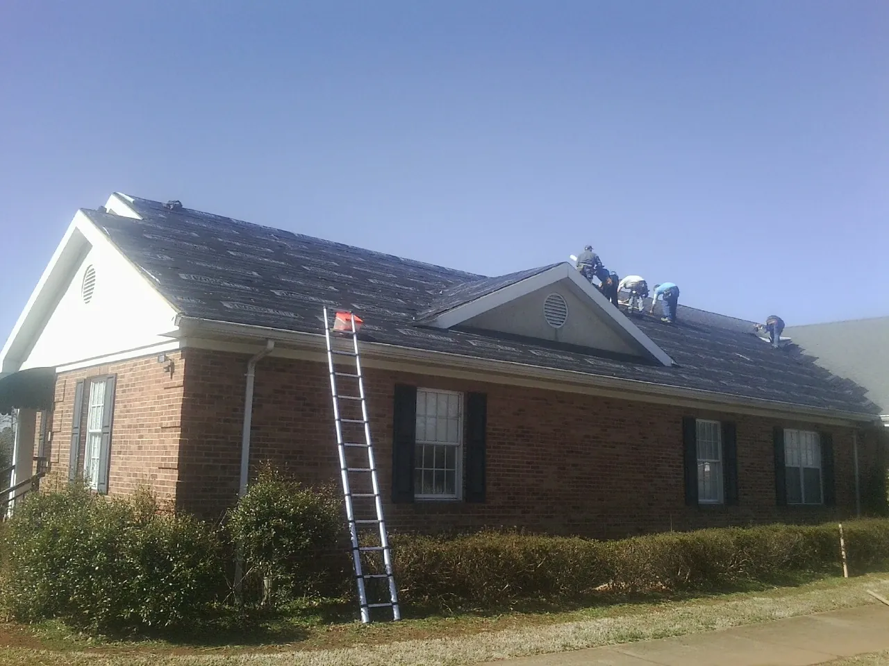 Crew tearing shingles off a brick ranch roof with a ladder set — Emergency Roof Repair work by our crew in New Smyrna Beach