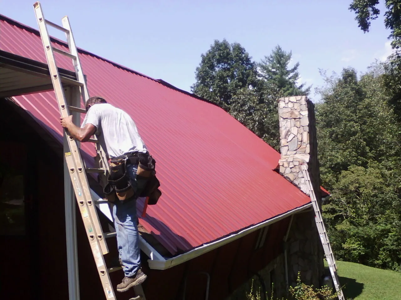Roofer on a ladder beside a red metal roof and stone chimney — Soffit & Fascia Repair by Roofing Brothers in New Smyrna Beach